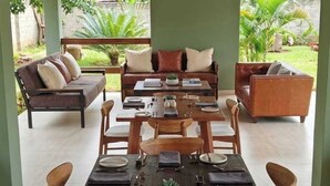 An outdoor dining area with a long table set for six, surrounded by wooden chairs, under a thatched roof. - TyCounty Boulevard Apartments (Bulanga)