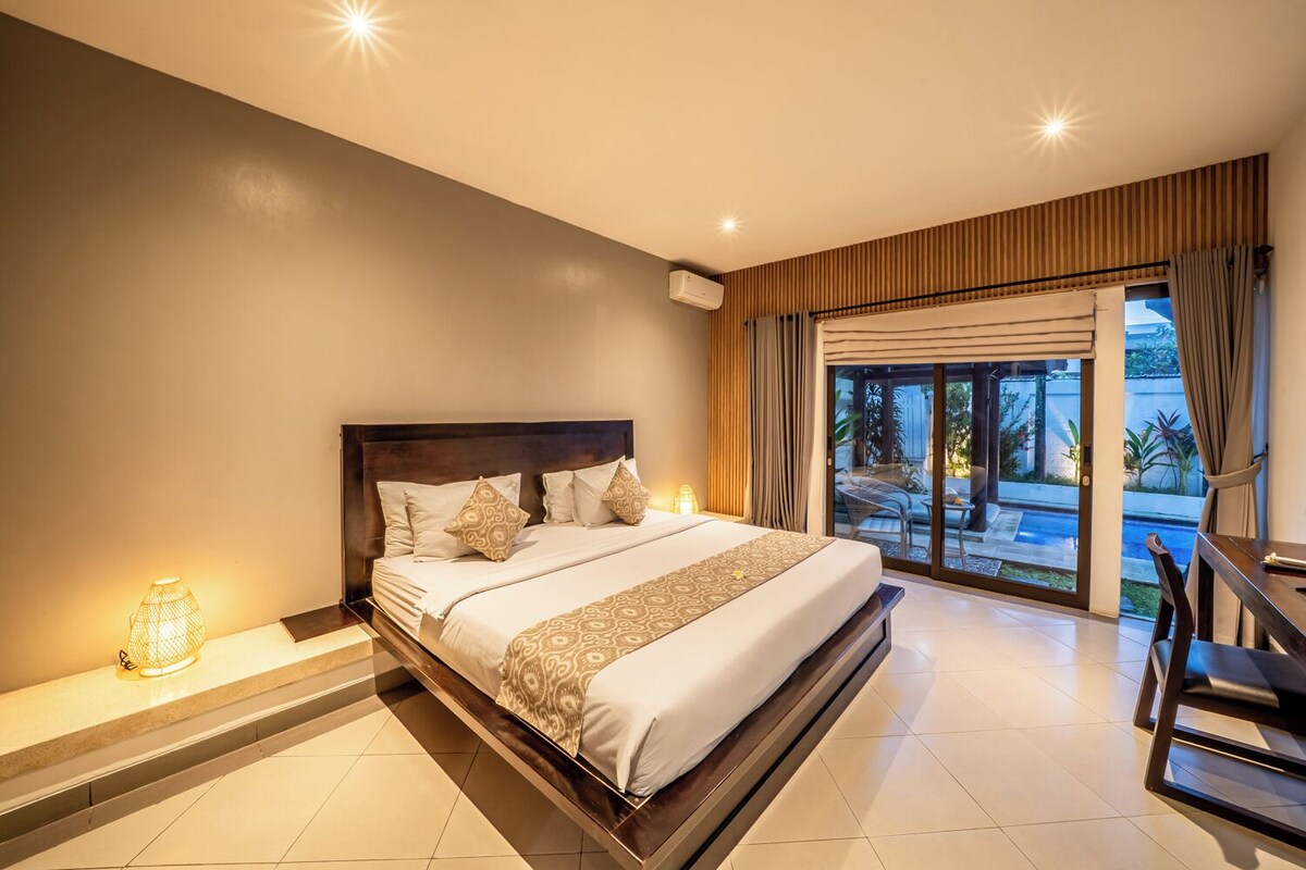 A hotel room with a large bed, a desk, a chair, a lamp, and a view of a pool and greenery.