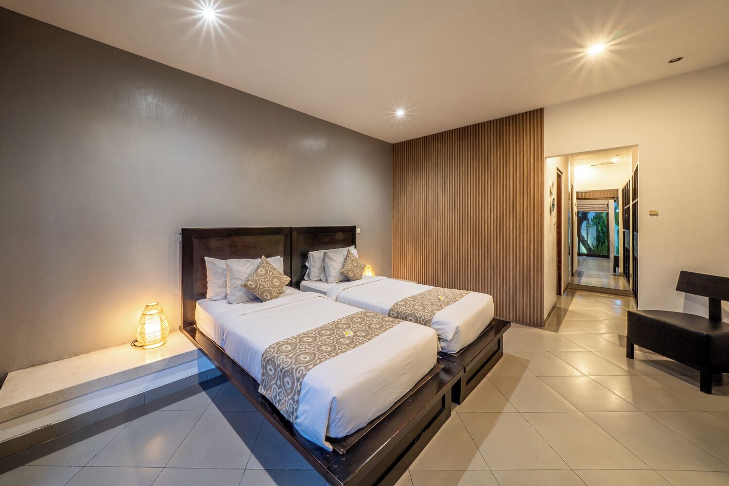 A hotel room with two beds, a bench, a lamp, and a view of a forest through a door.