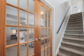A staircase with carpeted steps leading to a landing, a wooden-framed door with glass panels, and a view into a living room with a sofa and a lamp.