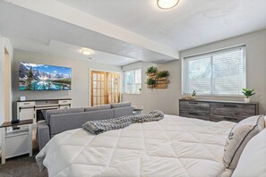 A bedroom with a large bed, a TV mounted on the wall, a sofa, a dresser, and a window with blinds.