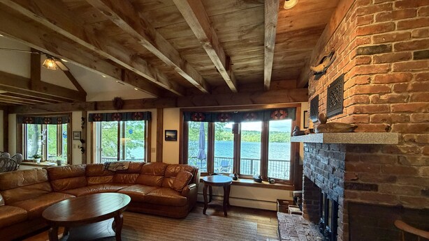 A cozy living room with a leather sectional sofa, a round coffee table, and a fireplace. Large windows offer views of the outdoors. - Wine'd Down on Lake Waukewan- New Lisiting- Photos Updated (Meredith)