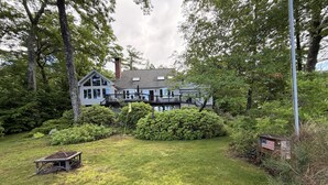 A two-story house with a deck, surrounded by lush greenery and a well-maintained lawn. - Wine'd Down on Lake Waukewan- New Lisiting- Photos Updated (Meredith)