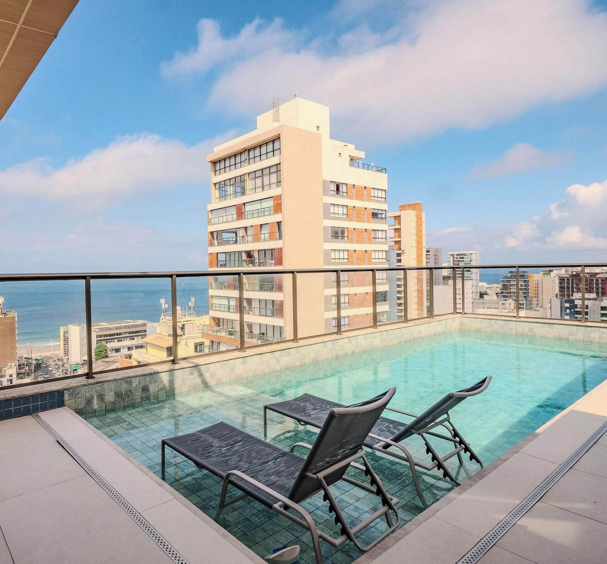 Una piscina al terrat amb dues cadira relaxants, vista a l'oceà i un edifici de pisos al fons.