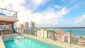 A rooftop swimming pool with a city view, a bar area, and a high-rise building in the background. - Relax in an ocean view apartment in Barra (Salvador)