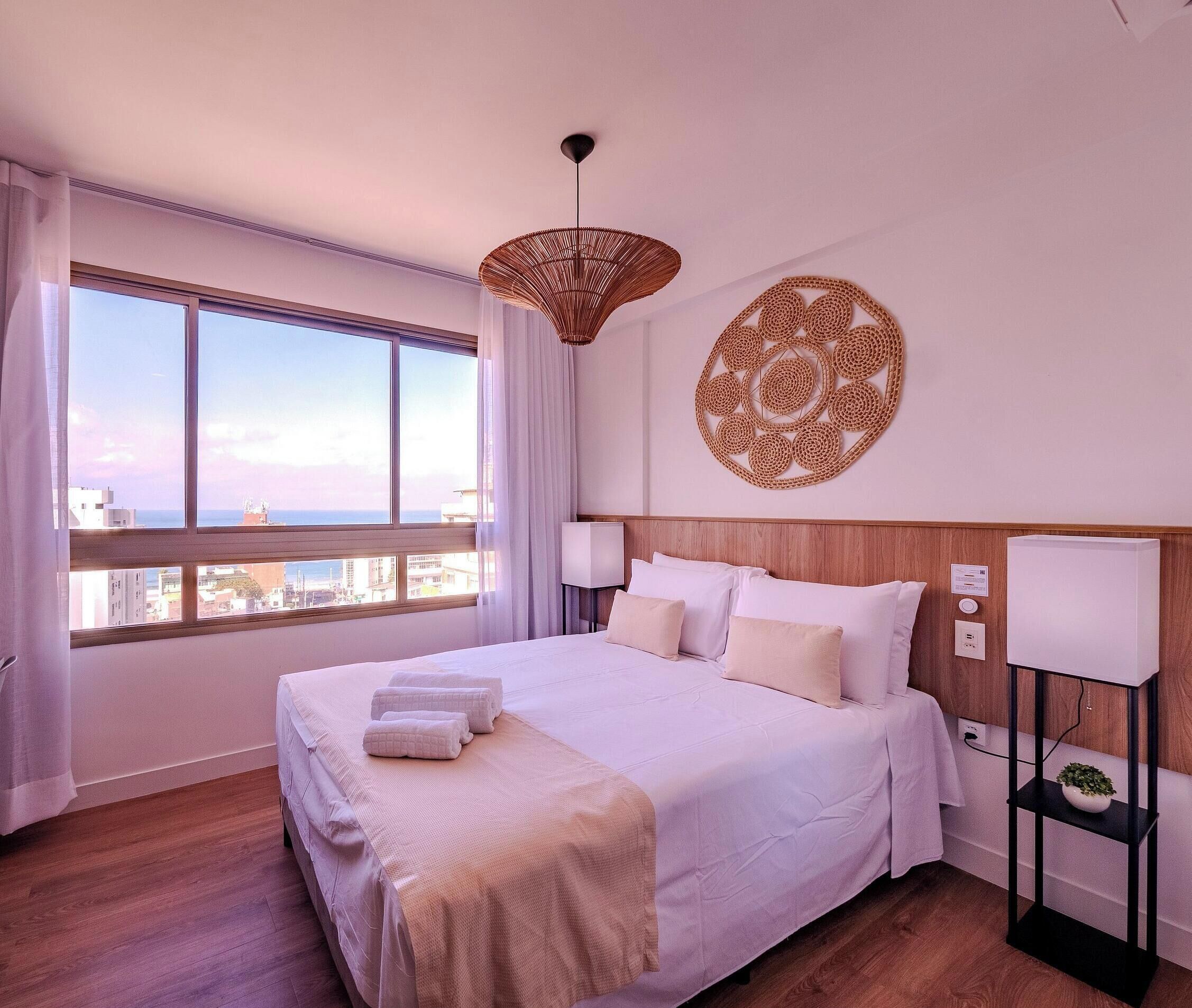 A hotel room with a large bed, a view of the city, a decorative wall piece, and a bedside table with a lamp.