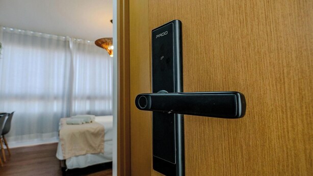 A modern door with a Pado lock mechanism, a wooden interior, and a glimpse of a bed and curtains in the background. - Relax in an ocean view apartment in Barra (Salvador)