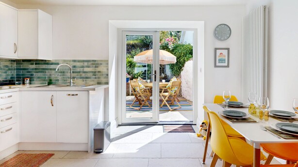 A modern kitchen with white cabinets, a tiled backsplash, and a sliding glass door leading to an outdoor dining area with yellow chairs and a table set for two.