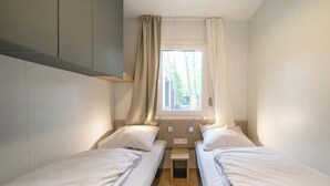Ett litet, kompakt rum med två enkelsängar, ett fönster med gardiner, ett litet bord och ett väggmonterat skåp.