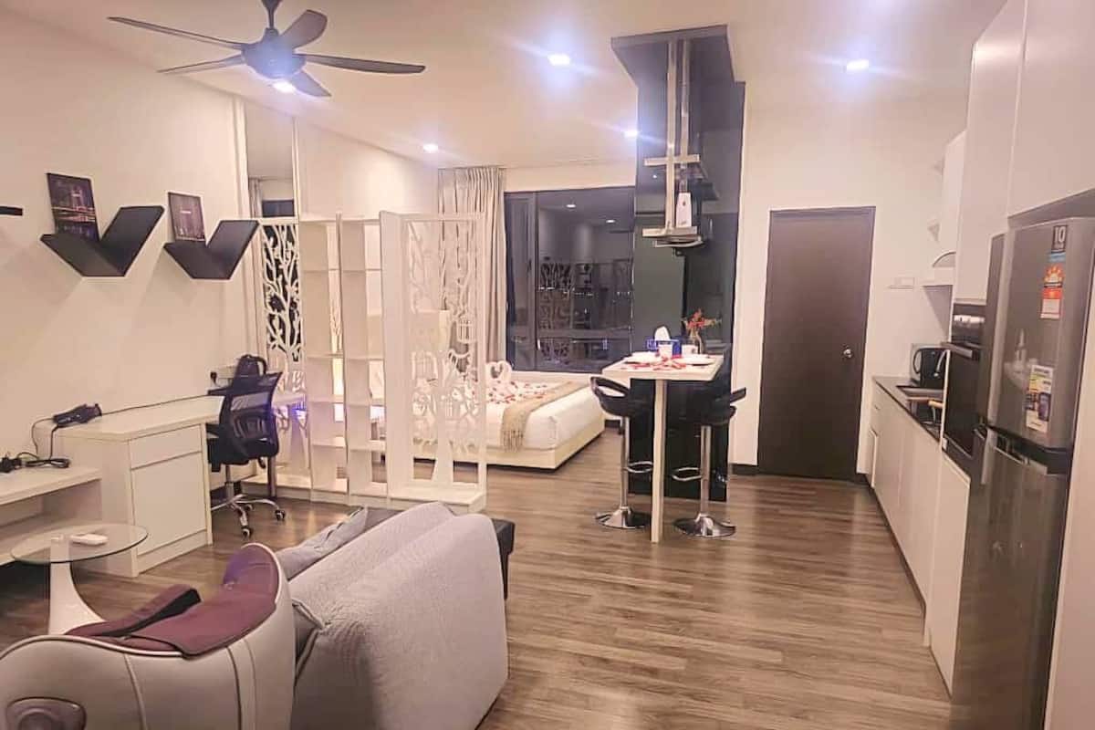 Uma sala de estar moderna com sofá, escrivaninha, poltrona, área de refeições com mesa e cadeiras, cozinha com geladeira e ventilador de teto.