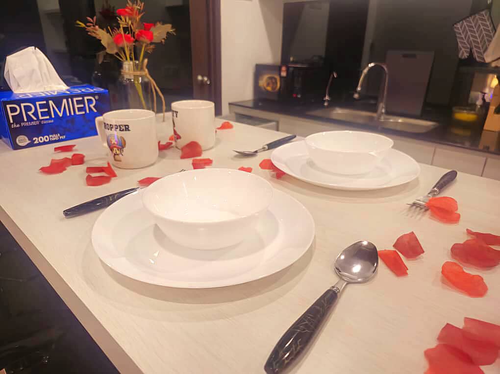 Een eettafel gedekt met witte borden, kommen, bestek en rozenblaadjes. Een doos zakdoekjes van Premier en een vaas met bloemen zijn ook te zien.