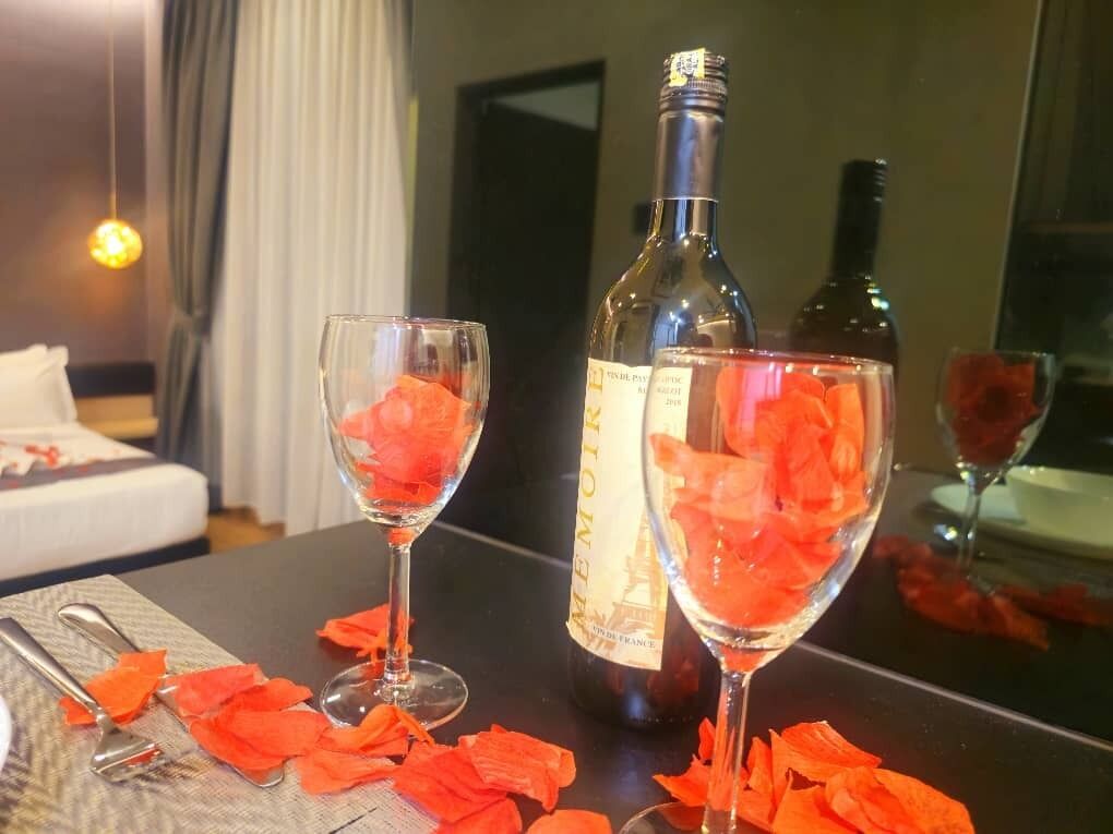 A room with a bed, a table with two glasses of a red drink, a bottle of wine, and scattered rose petals.
