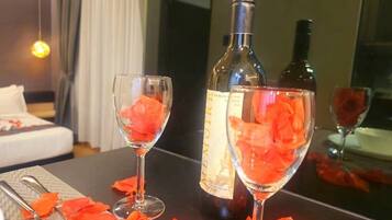 A room with a bed, a table with two glasses of a red drink, a bottle of wine, and scattered rose petals.