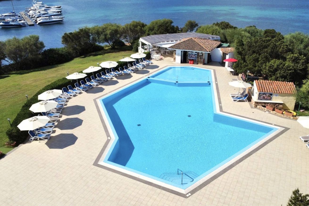 A swimming pool with lounge chairs and umbrellas, a small building, and boats in the background.