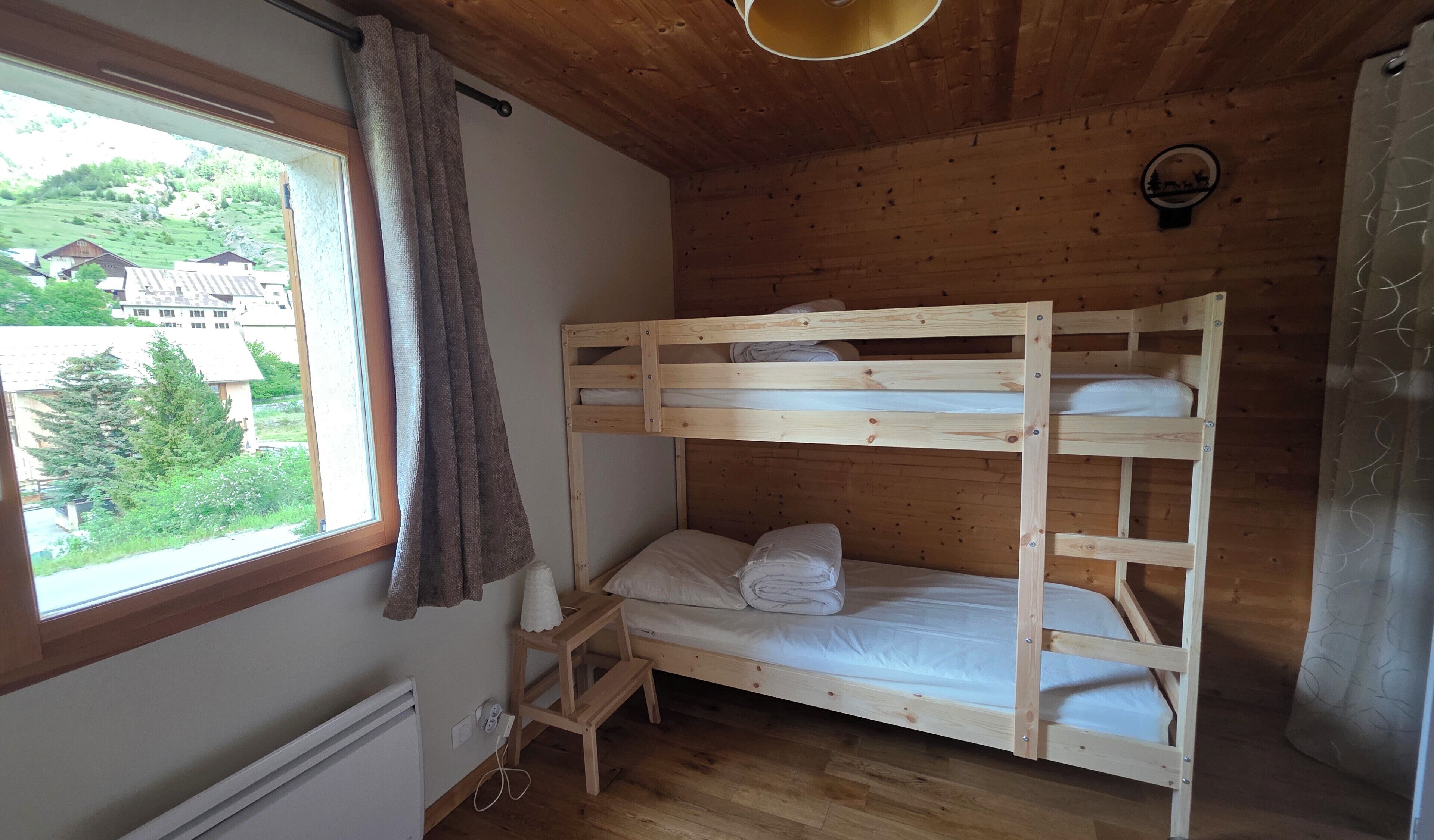 Una cama litera con estructura de madera, una ventana con cortinas, un taburete pequeño de madera y un radiador.