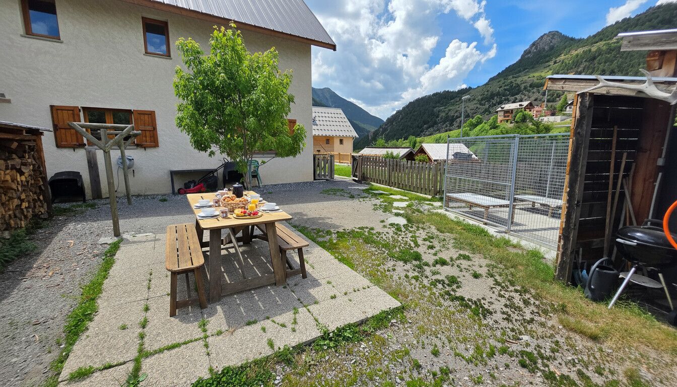 A rustic outdoor dining area with a table and benches, a wooden pergola, and a mountain backdrop.