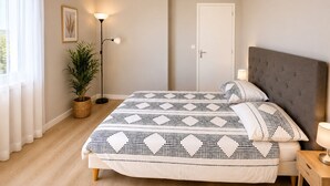 Un dormitorio con cama, mesita de noche, lámpara, planta en maceta y una puerta.