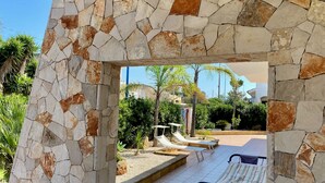 A stone archway leading to a pool area with lounge chairs and palm trees. - Villa Adua | Blueapulia (Torre Suda)