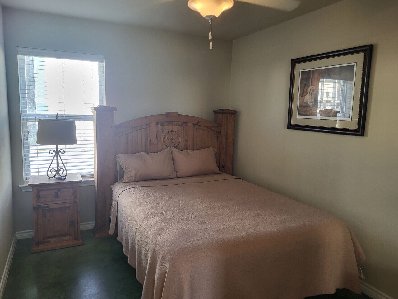 A bedroom with a bed, a nightstand, a lamp, a framed picture on the wall, and a window with blinds.