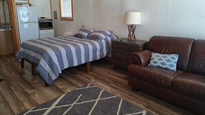 A bedroom with a bed, a brown leather sofa, a nightstand, a lamp, and a kitchen area with a refrigerator and microwave.