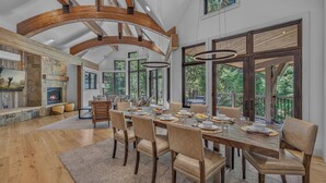 Une salle à manger spacieuse avec un plafond haut, des poutres en bois et de grandes fenêtres offrant une vue sur l’extérieur.