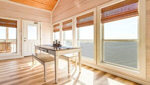 Um quarto com teto de madeira, grandes janelas com persianas, uma mesa de madeira com um banco e vista para a água.