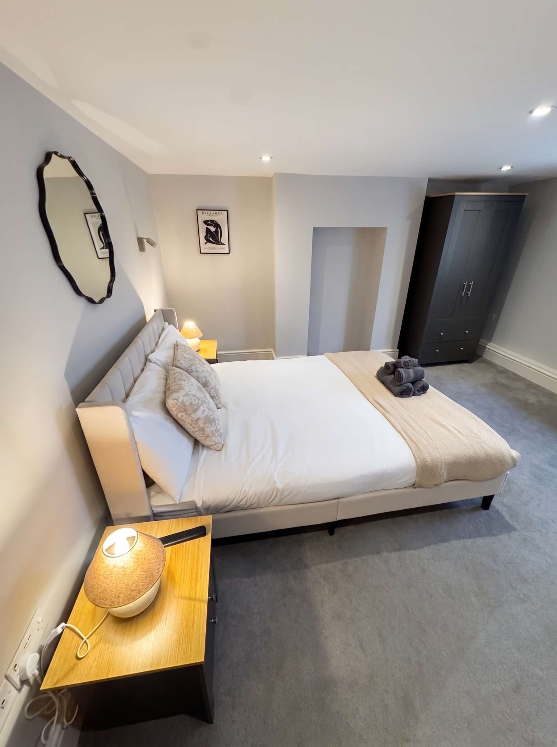Une chambre moderne avec un grand lit, des lampes de chevet, une petite table avec une lampe, une armoire et un miroir fixé au mur.