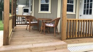 A covered porch with wooden railings, two wicker chairs, a small table, and a white RV parked in the background. - Tiny Home at the Beach (Port O`Connor)