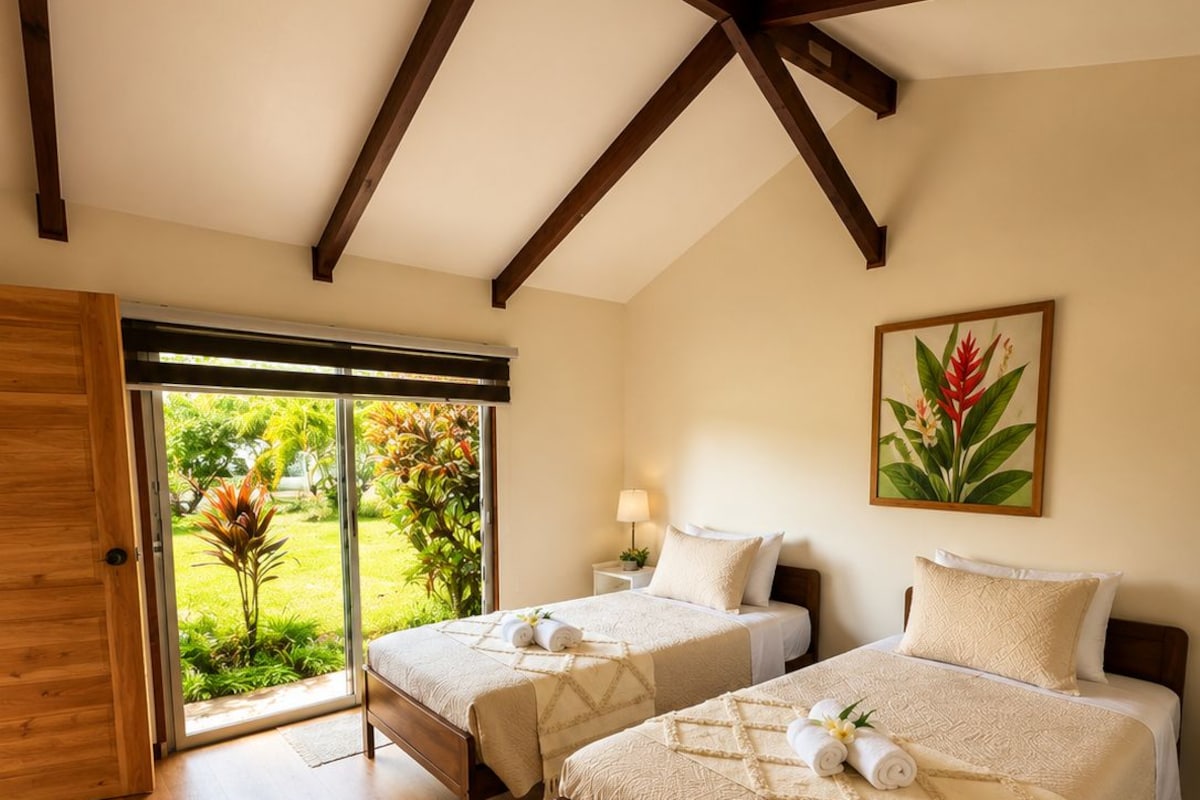 A bedroom with two beds, wooden beams, a large window, and a tropical plant.