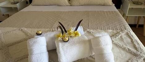 Una cama bien tendida con colcha, dos toallas con flores decorativas y lámparas de noche sobre una mesita blanca.