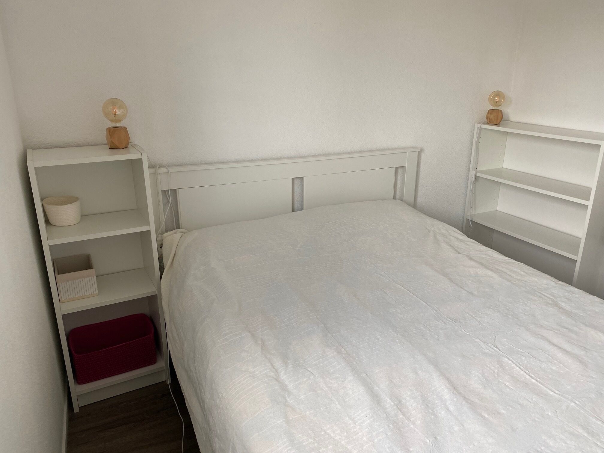 A bedroom with a white bed, a wooden headboard, and a white shelving unit with a red cushion.