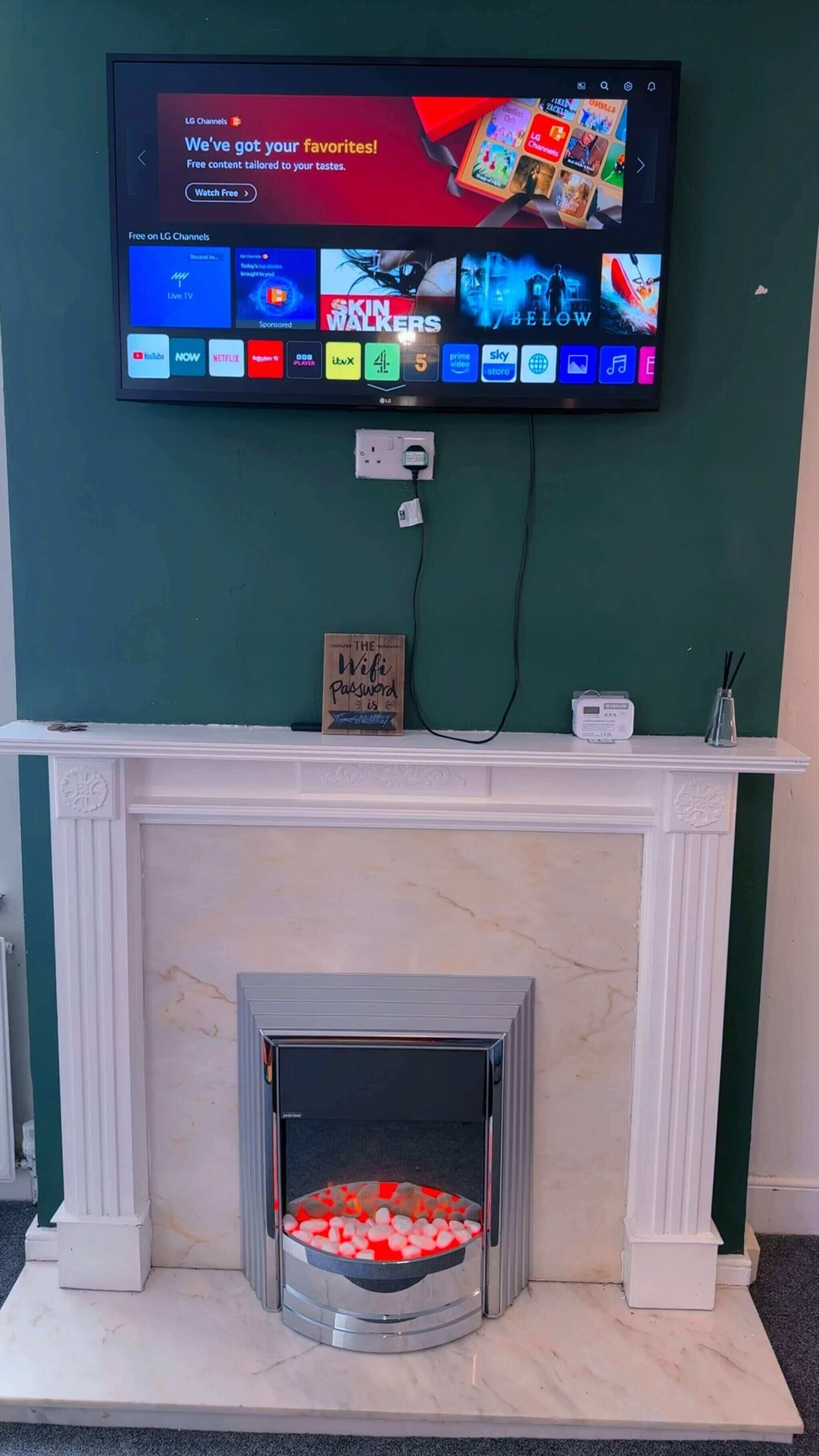A television mounted on a wall displaying various channels and content, a fireplace with a decorative sign, and a power outlet below.