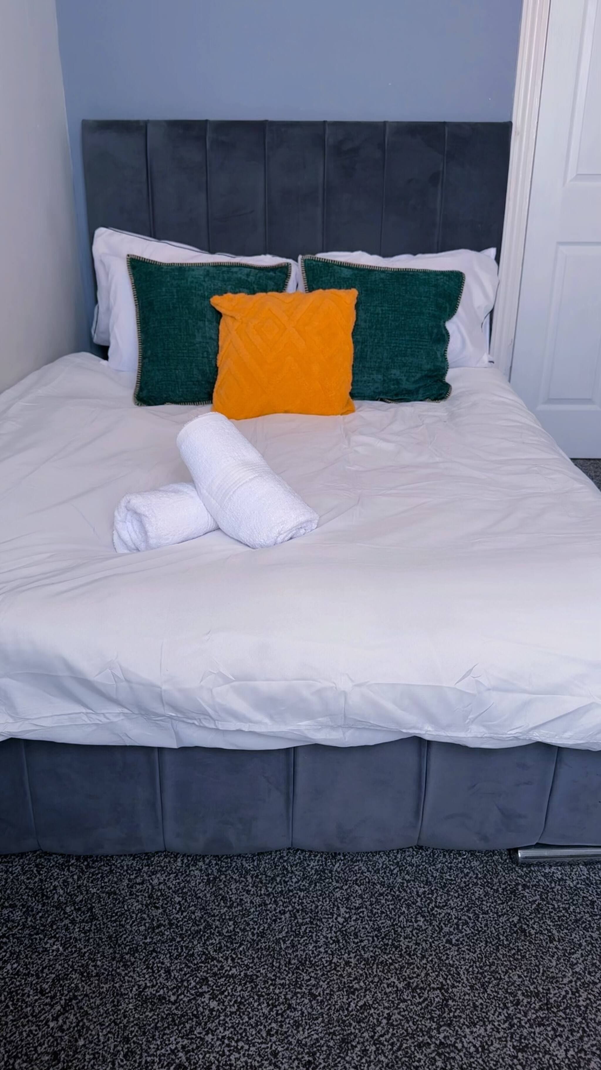 Una cama bien hecha con un edredón blanco, dos almohadas verdes y una almohada naranja. Una toalla blanca doblada está al pie de la cama.