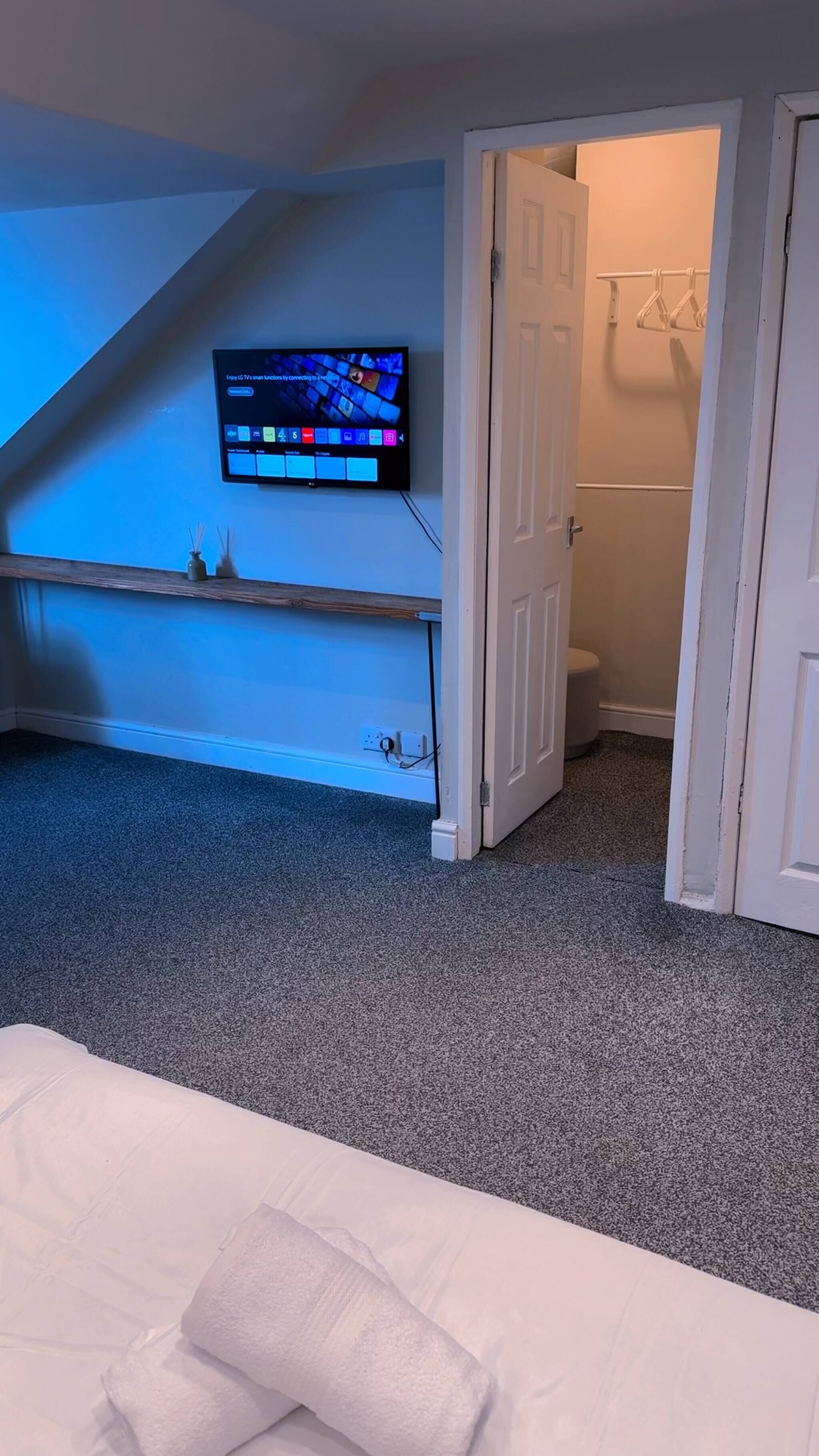 Une chambre avec un téléviseur fixé au mur, une porte donnant sur une salle de bain et un lit blanc avec un oreiller.