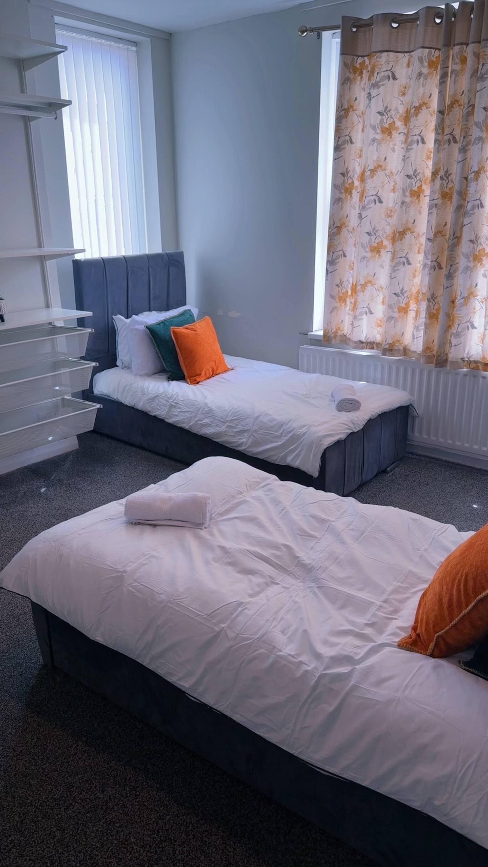 A bedroom with two beds, a window with blinds, a radiator, and a curtain.