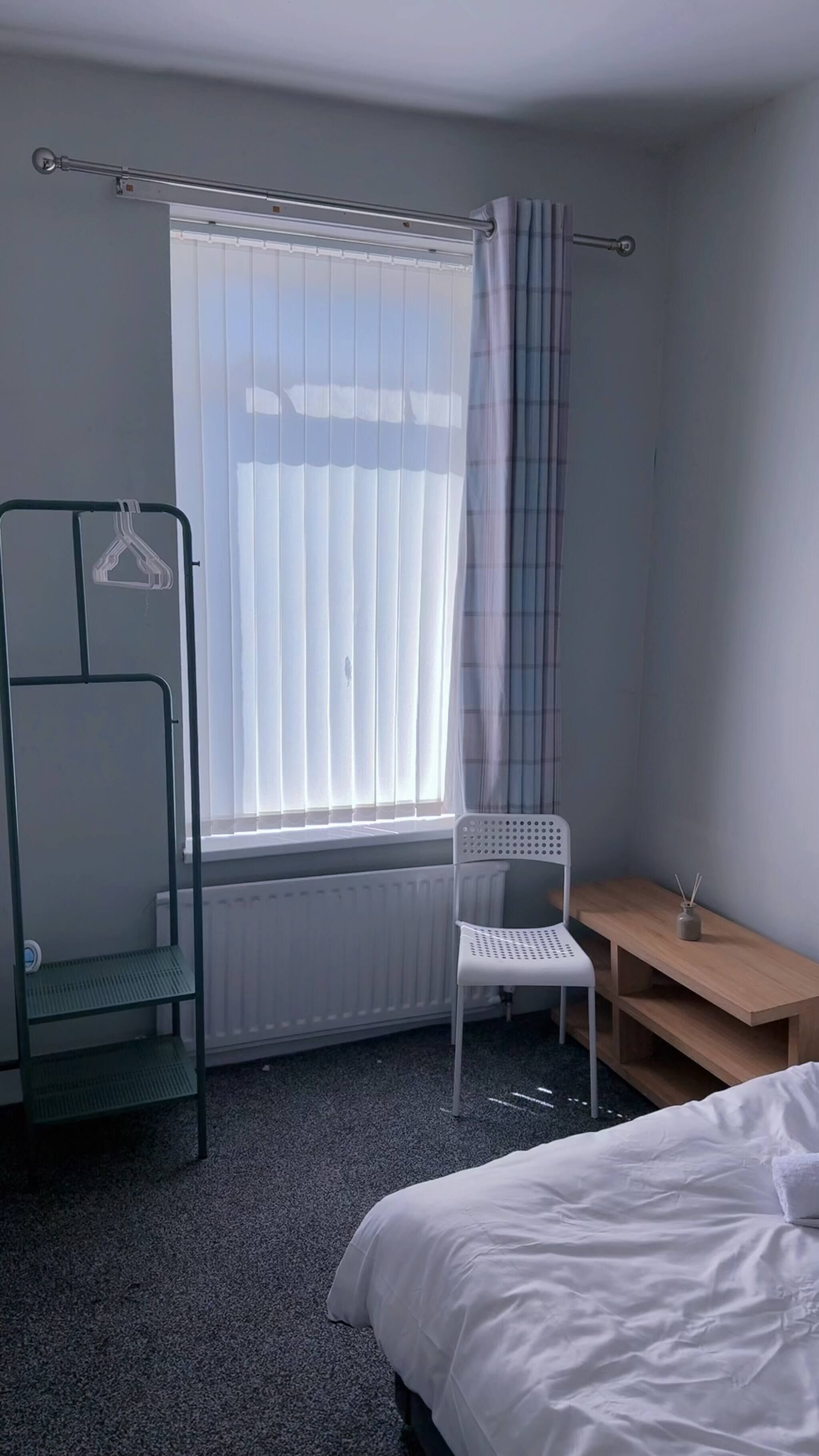 A bedroom with a bed, a chair, a radiator, a window with curtains, and a shelf.