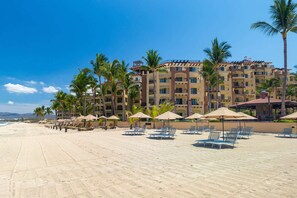 Ein Hotel am Strand mit mehreren Balkonen, Liegestühlen und Palmen.