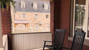 Un balcón con piso de madera, dos mecedoras, una ventana y vista a un edificio con cerca blanca.