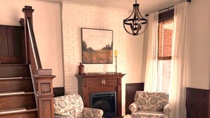 Una sala de estar con chimenea, dos sillones con estampado, una escalera de madera y un candelabro.