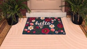 Puerta principal con placa de identificación, felpudo con la palabra 'hello' y motivos florales, y dos macetas con plantas a cada lado.