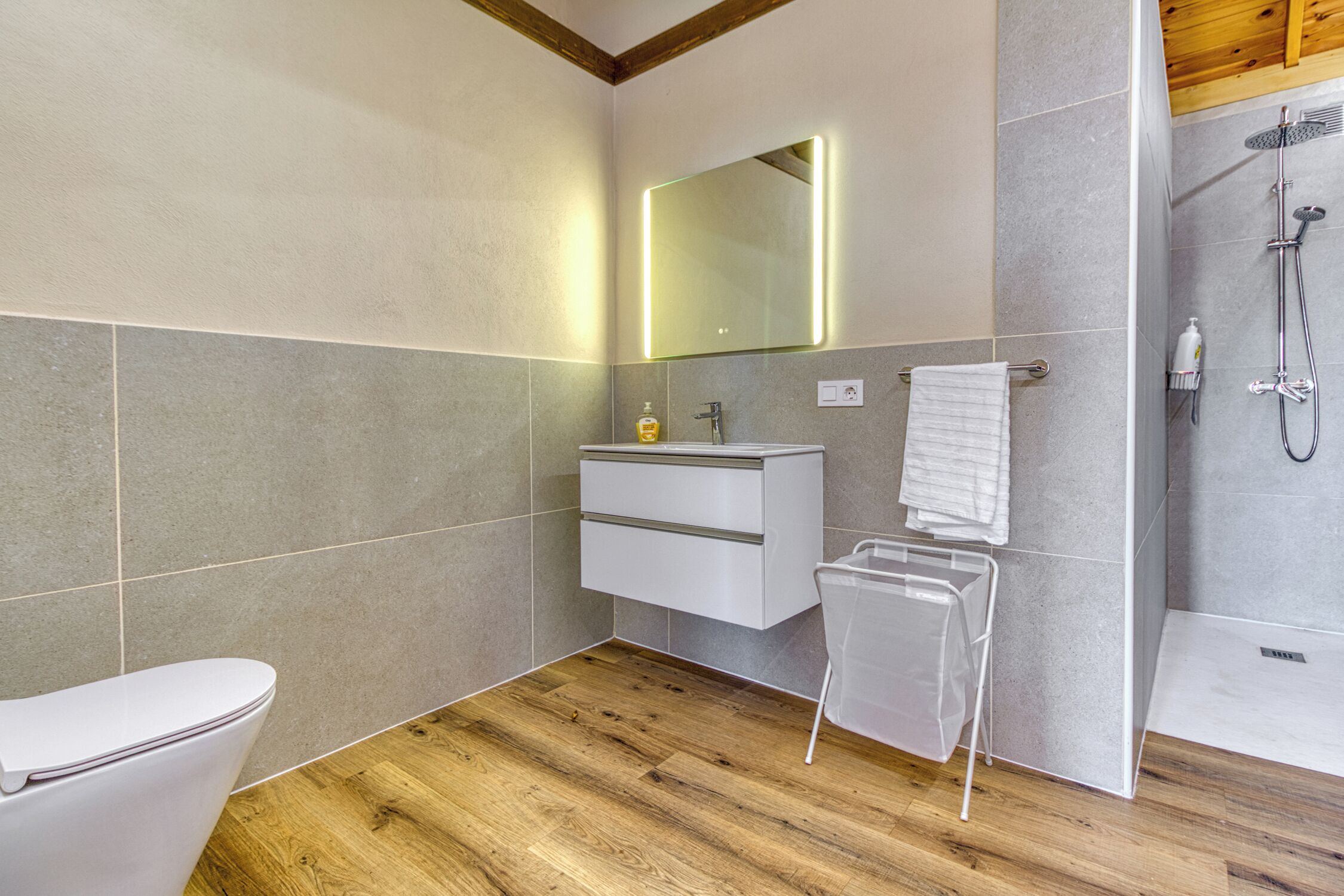 Ein modernes Badezimmer mit Holzboden, Toilette in Weiß, Waschbecken mit Spiegel und Duschbereich.
