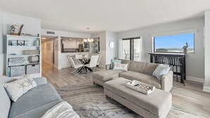 A modern living room with a sectional sofa, a dining area, and a kitchen in the background. - 😎🏄🌞Bright and Sunny Oceanfront | Seascape Towers 425🏖️🌞🐳 (New Smyrna Beach)