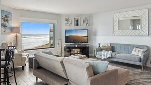 A living room with a large TV, a sofa, a dining area, and a view of the beach. - 😎🏄🌞Bright and Sunny Oceanfront | Seascape Towers 425🏖️🌞🐳 (New Smyrna Beach)