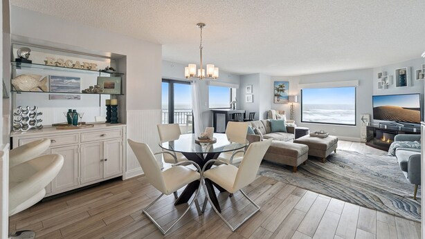 A modern living room with a glass dining table, white sofas, a TV, and a large window with a view. - 😎🏄🌞Bright and Sunny Oceanfront | Seascape Towers 425🏖️🌞🐳 (New Smyrna Beach)
