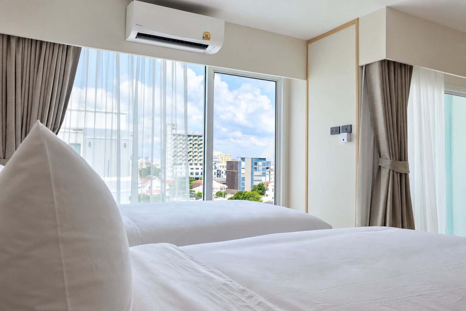 A hotel room with two beds, an air conditioning unit, a window with curtains, and a view of buildings outside.