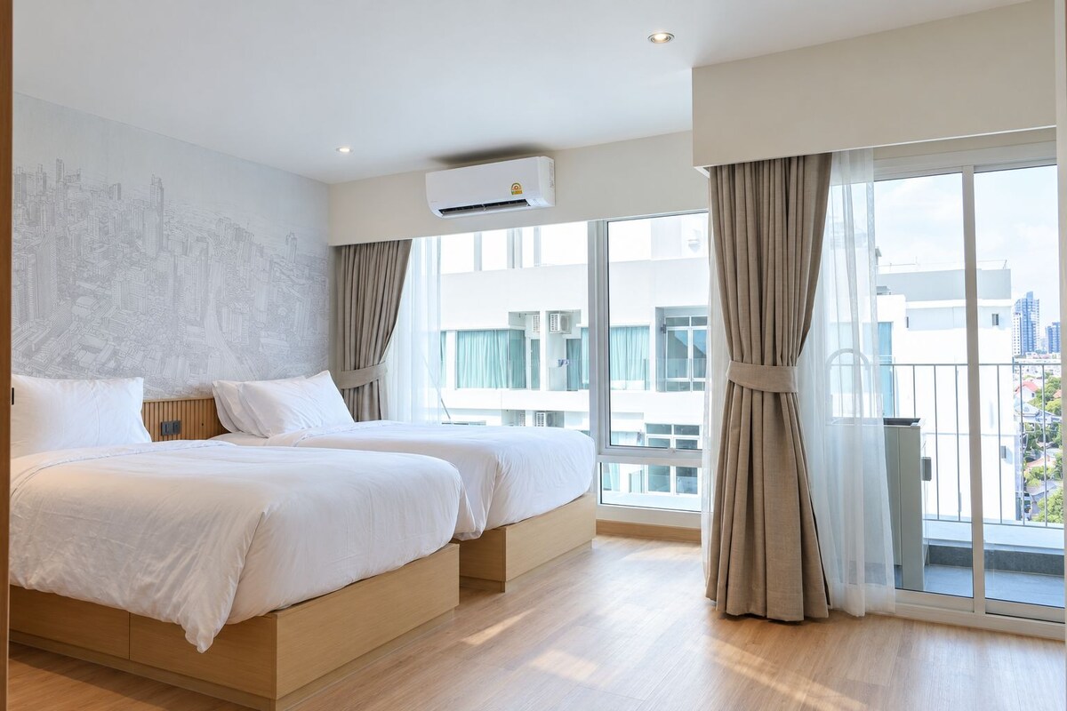 Una habitación de hotel moderna con dos camas, un mural con un paisaje urbano, ventanas amplias y un equipo de aire acondicionado.