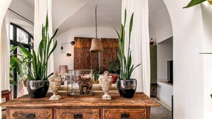 Una mesa de madera con jarrones decorativos y una vitrina de vidrio, bordeada por altas plantas.