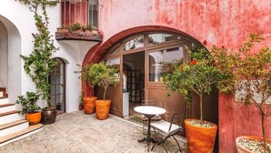 Un patio con un edificio blanco, un muro rojo, un balcón con plantas y una mesa con sillas.