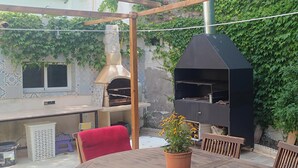 Un patio al aire libre con mesa y sillas de madera, una silla roja, una planta en maceta, una parrilla y una pared de baldosas cubierta de hiedra.