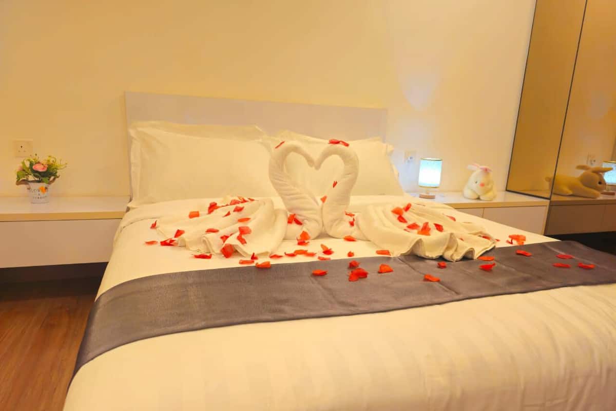 A hotel room with a neatly made bed, a heart-shaped towel arrangement, and a bedside table with a lamp and a small plant.
